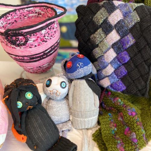  Various work of fibre art including three sock puppets, a woven basket, and a crocheted scarf.