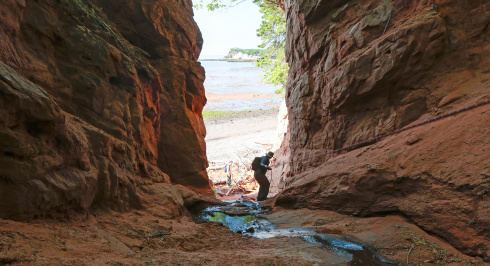 Two people wearing hats and backpacks navigate through a narrow, rocky crevasse toward a beach. The towering cliffs on either side are made of layered, reddish-brown sandstone. A small stream of water flows over the rocky ground in the foreground, leading toward the sandy shore and the blue waters of the Bay of Fundy in the distance under a clear sky.